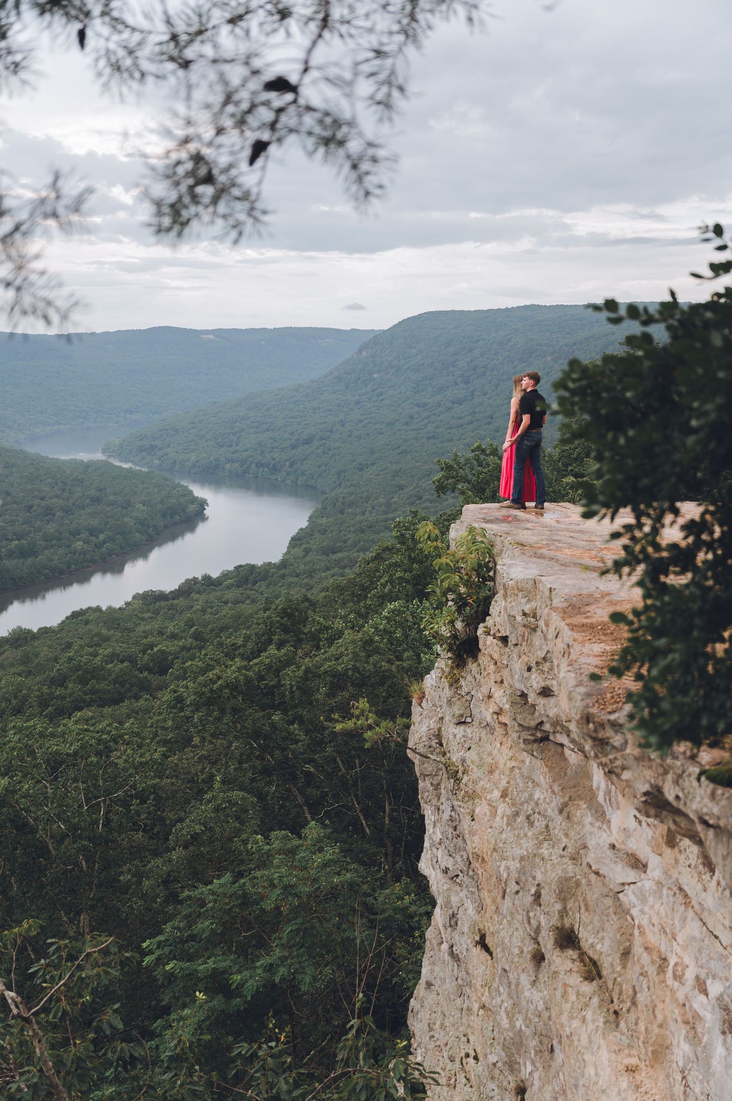 Two figures embrace on a dramatic cliff edge overlooking a winding river valley and lush forested mountains.