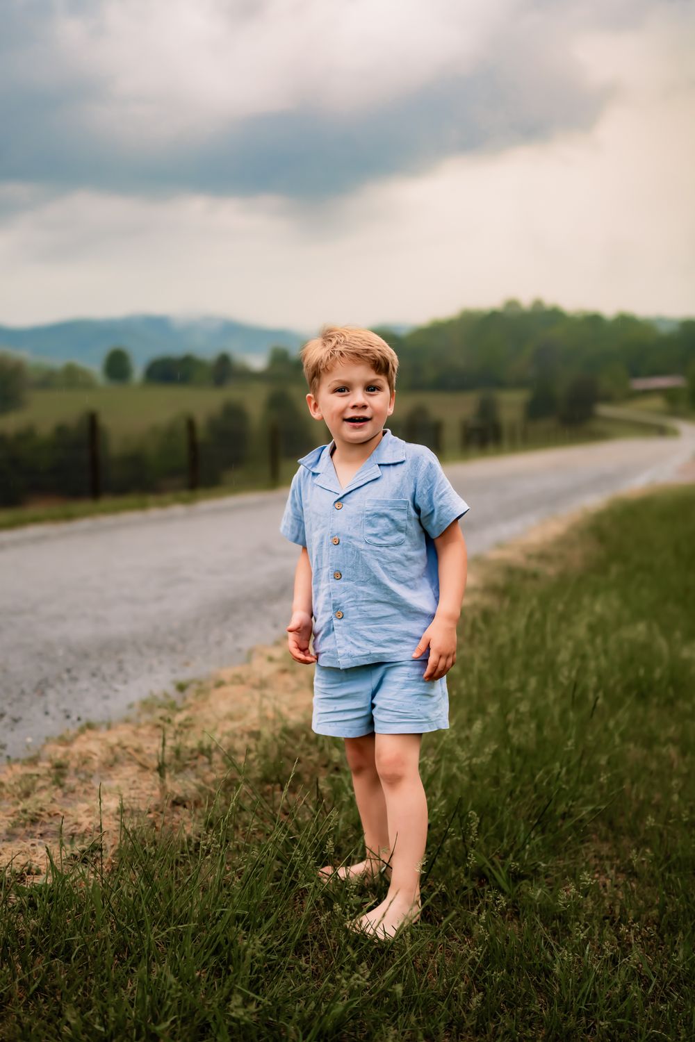 A small child in a blue outfit walks along a grassy path with mountains and cloudy skies in the background.