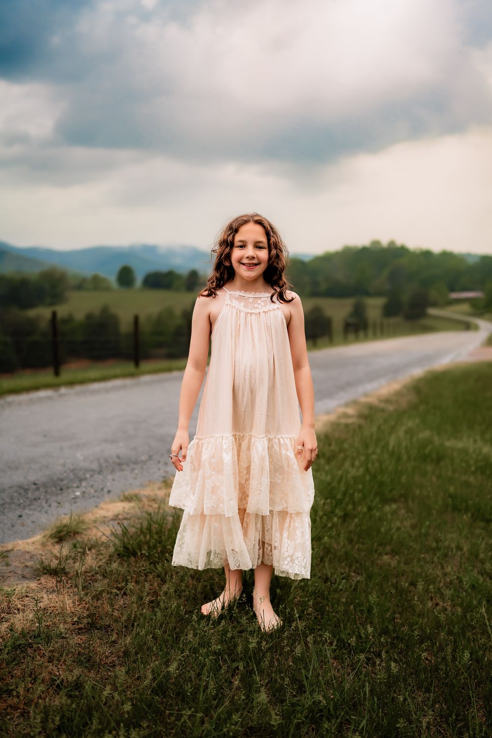 A child in a flowing cream dress sits near a curving country road on a cloudy summer day.