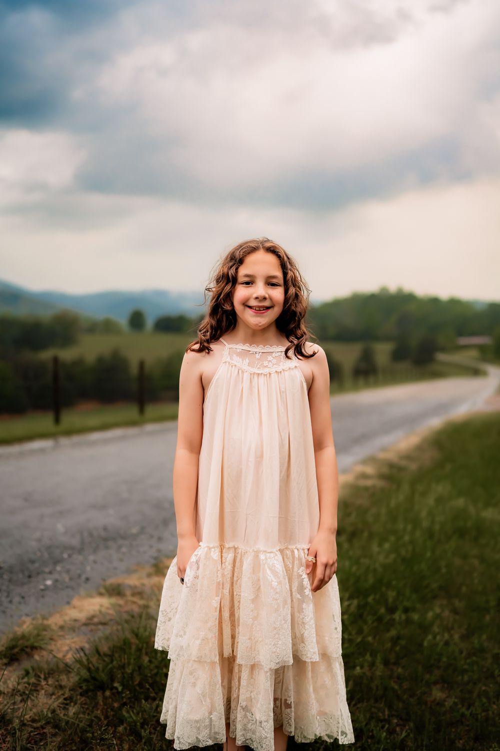 A young girl in a white lace dress stands in a field against a cloudy mountain landscape.