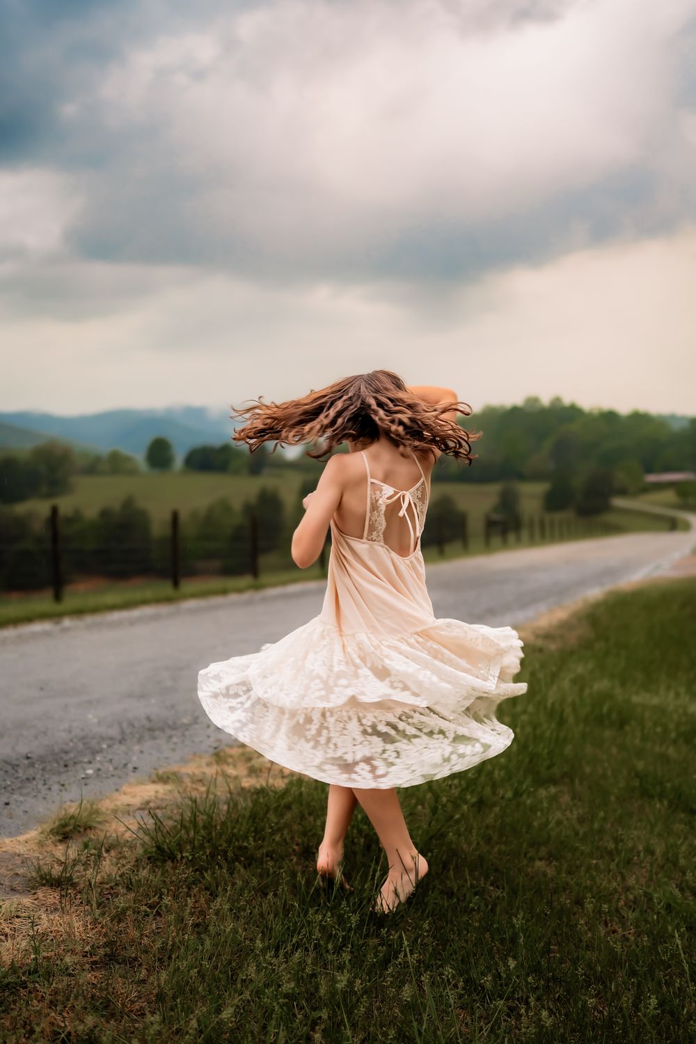 A figure in a flowing white dress twirls gracefully in a grassy field with mountains and stormy skies in the background.