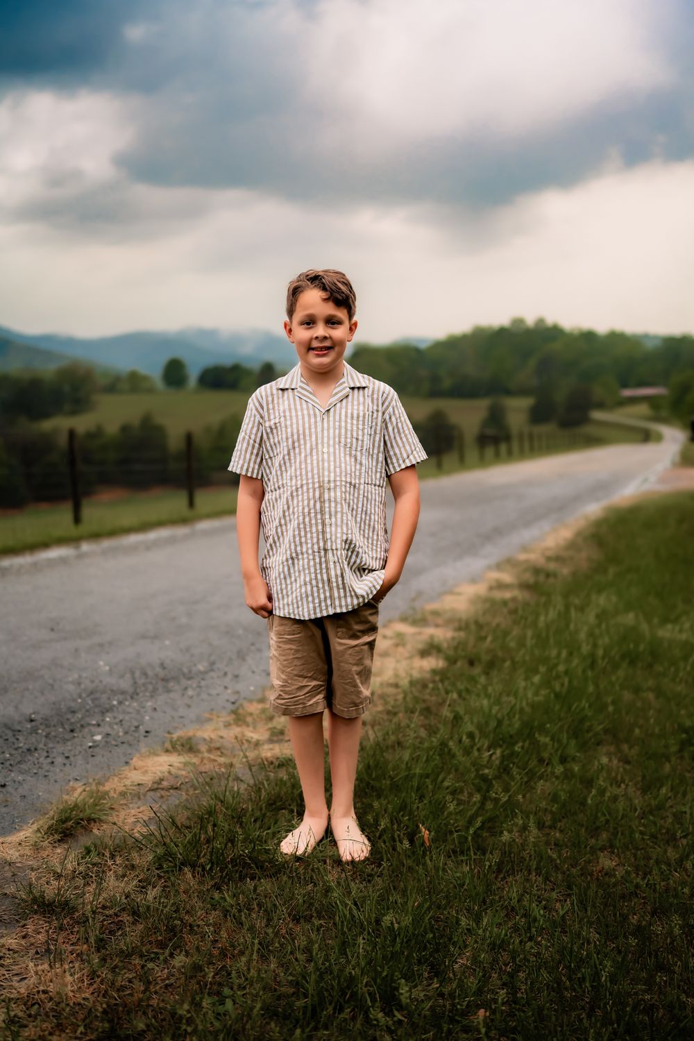Child standing on a grassy hillside along a winding country road at dusk wearing striped shirt and shorts.