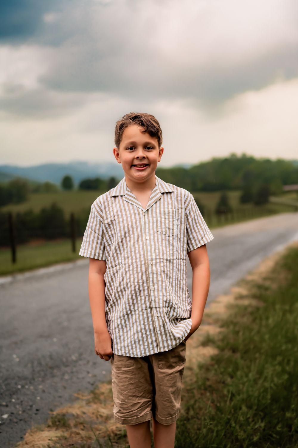 A young child in a striped shirt stands smiling on a rural mountain road on an overcast day.