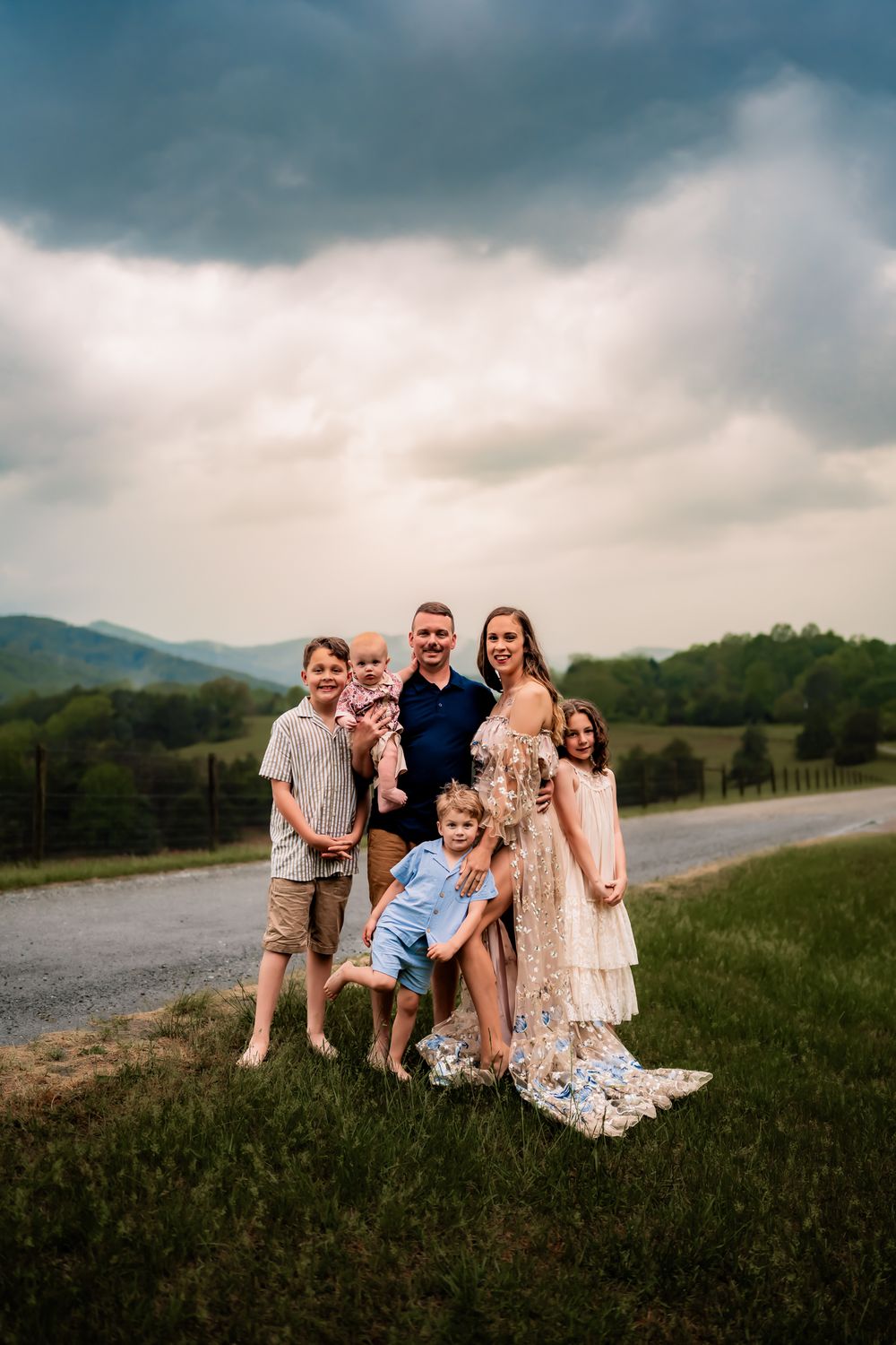 A family sits together on a grassy hill against a dramatic cloudy sky and mountain landscape.