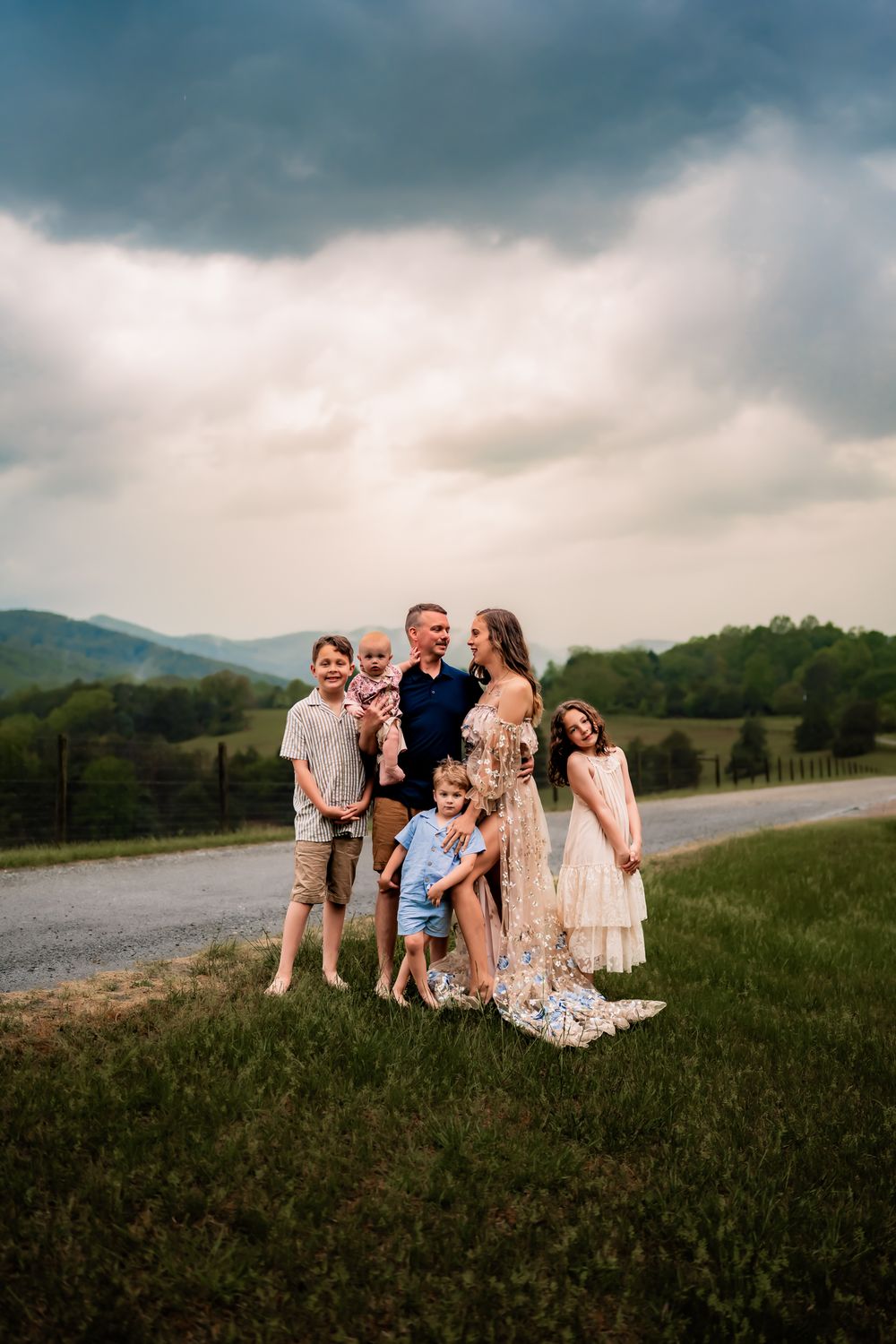 A family stands together on a grassy hillside with mountains and stormy clouds in the background at sunset.