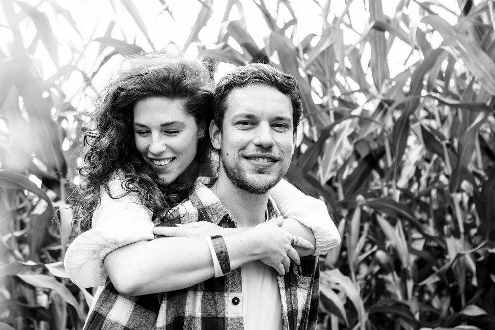 Vrouw sprint op de rug van haar vriend met houthakkers shirt. Ze staan in het maisveld. Zij houdt hem stevig vast. De foto is zwart wit