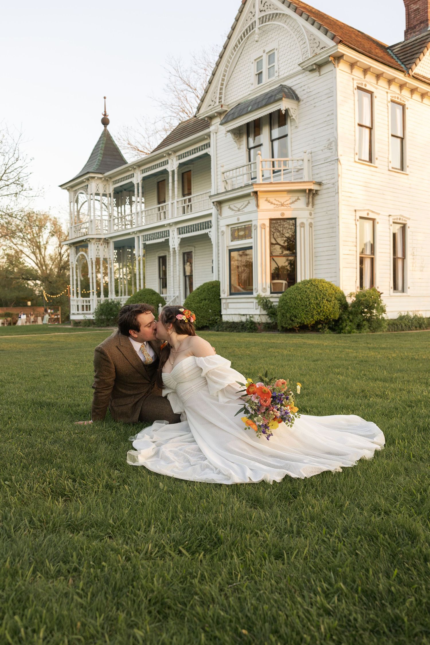 Couple in wedding attire sharing romantic moment on lawn of elegant Victorian mansion at sunset.
