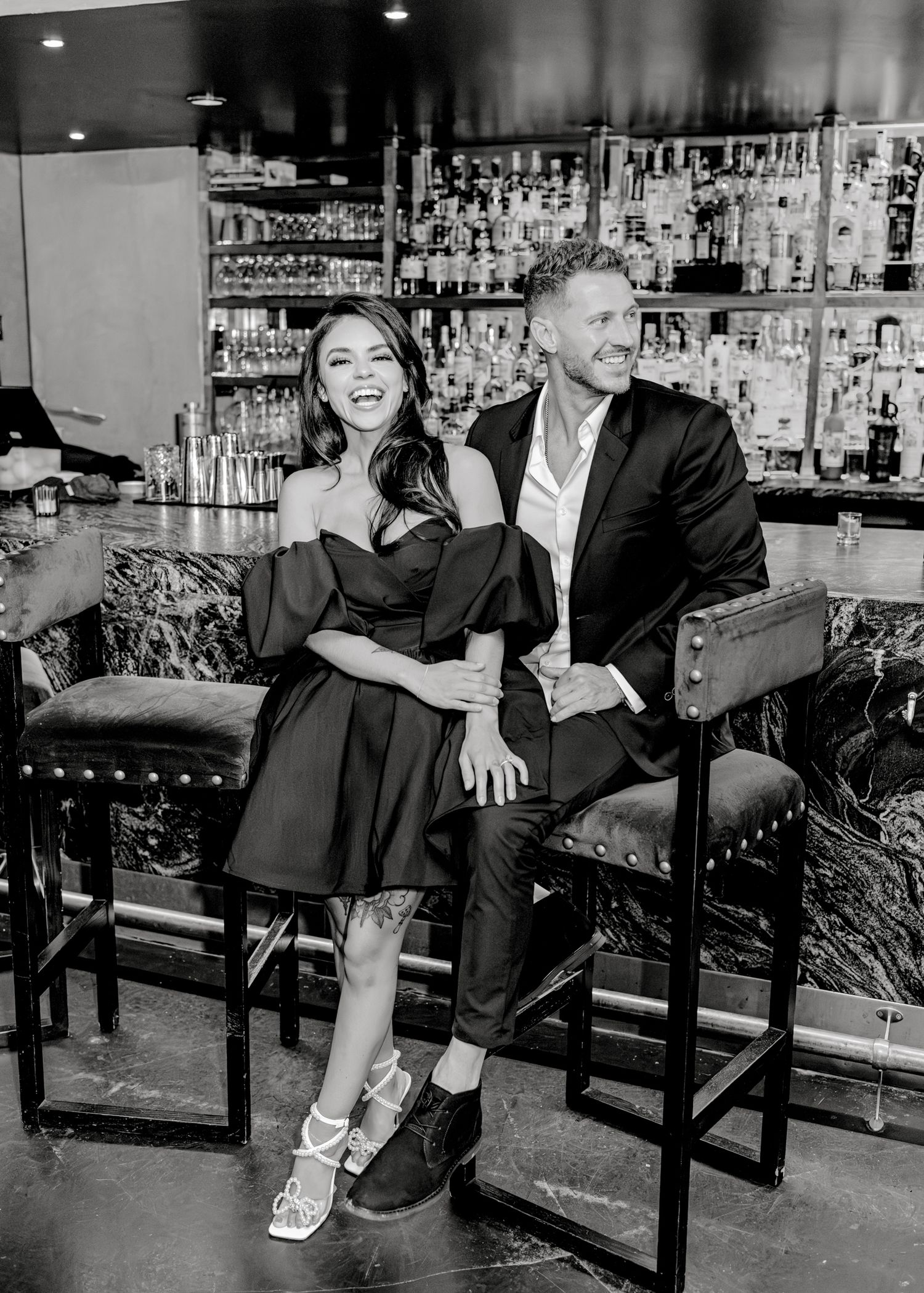 A black and white photo of a couple laughing together while seated at an elegant bar with a wall of liquor bottles behind them.