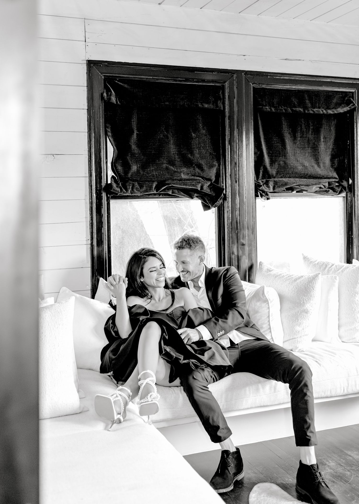 Black and white photo of a couple relaxing together on a white couch by a window with dark curtains.