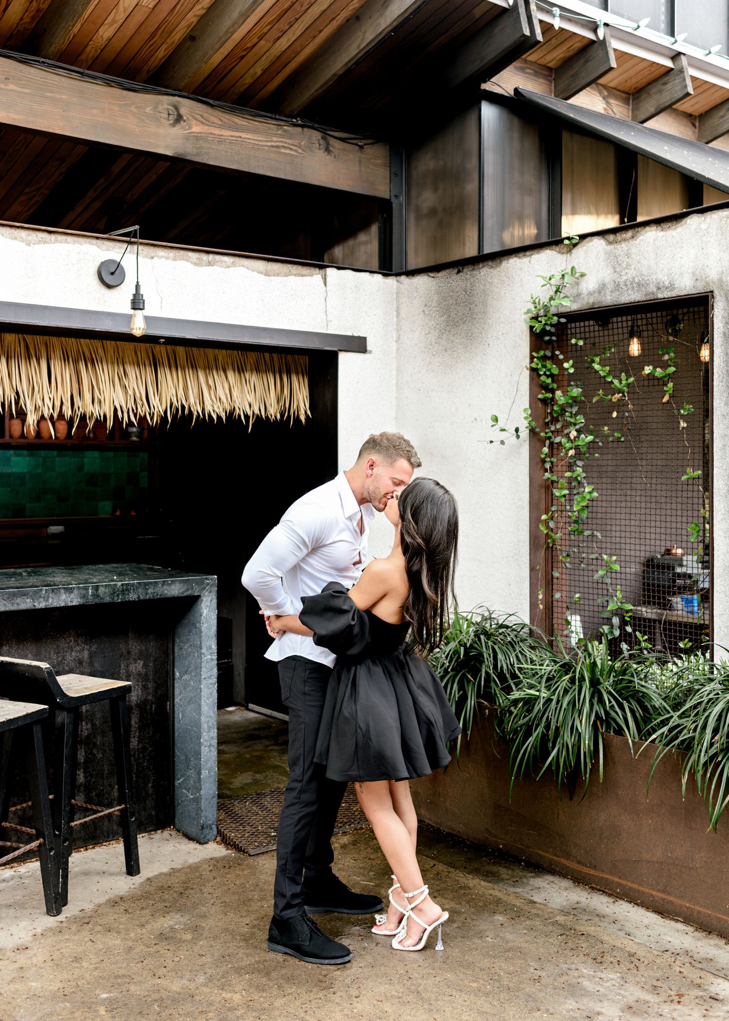 Couple in formal attire share an intimate moment in an outdoor covered patio area with tropical plants.