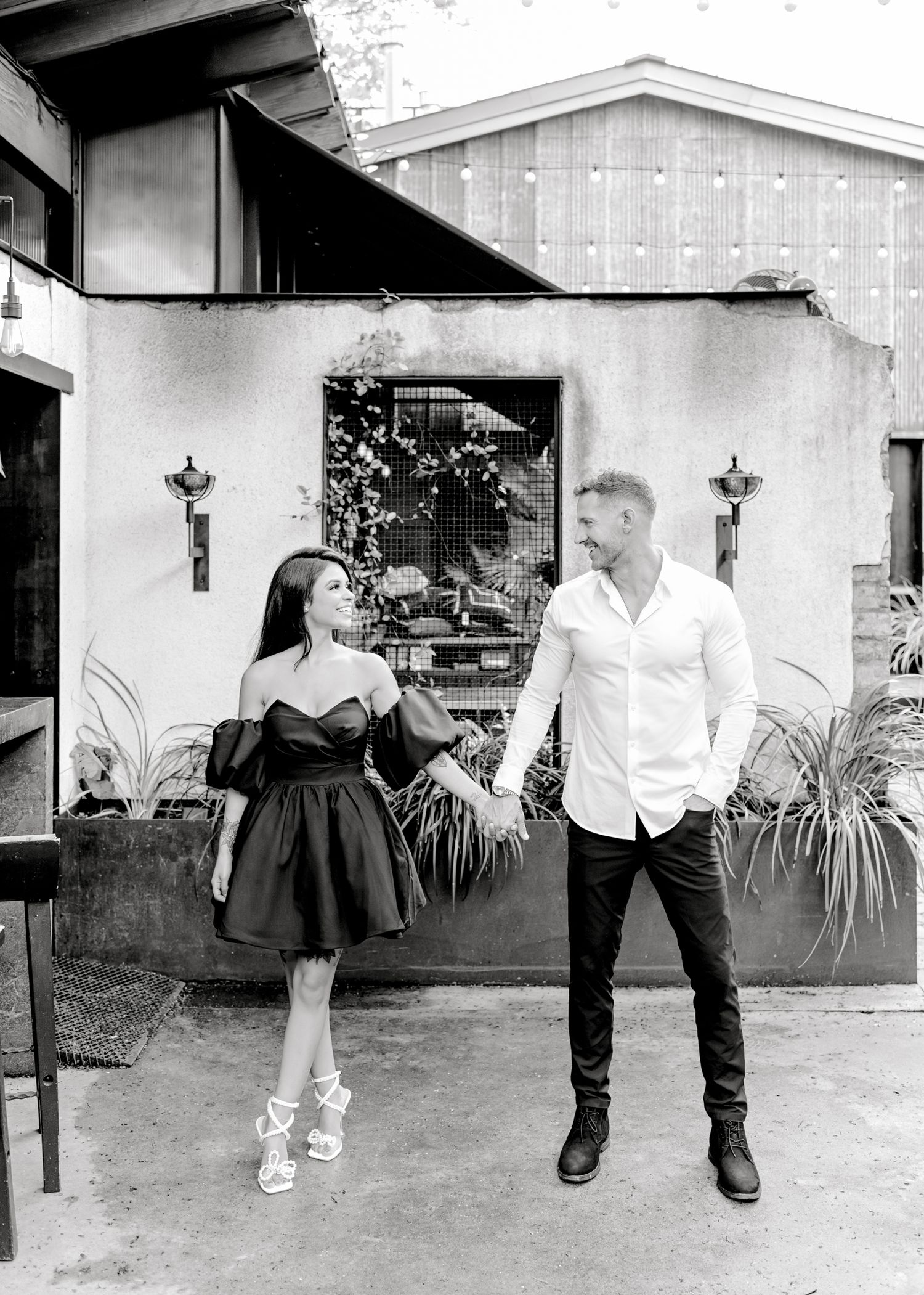 Black and white photo of couple posing against rustic wall with plants and lanterns.