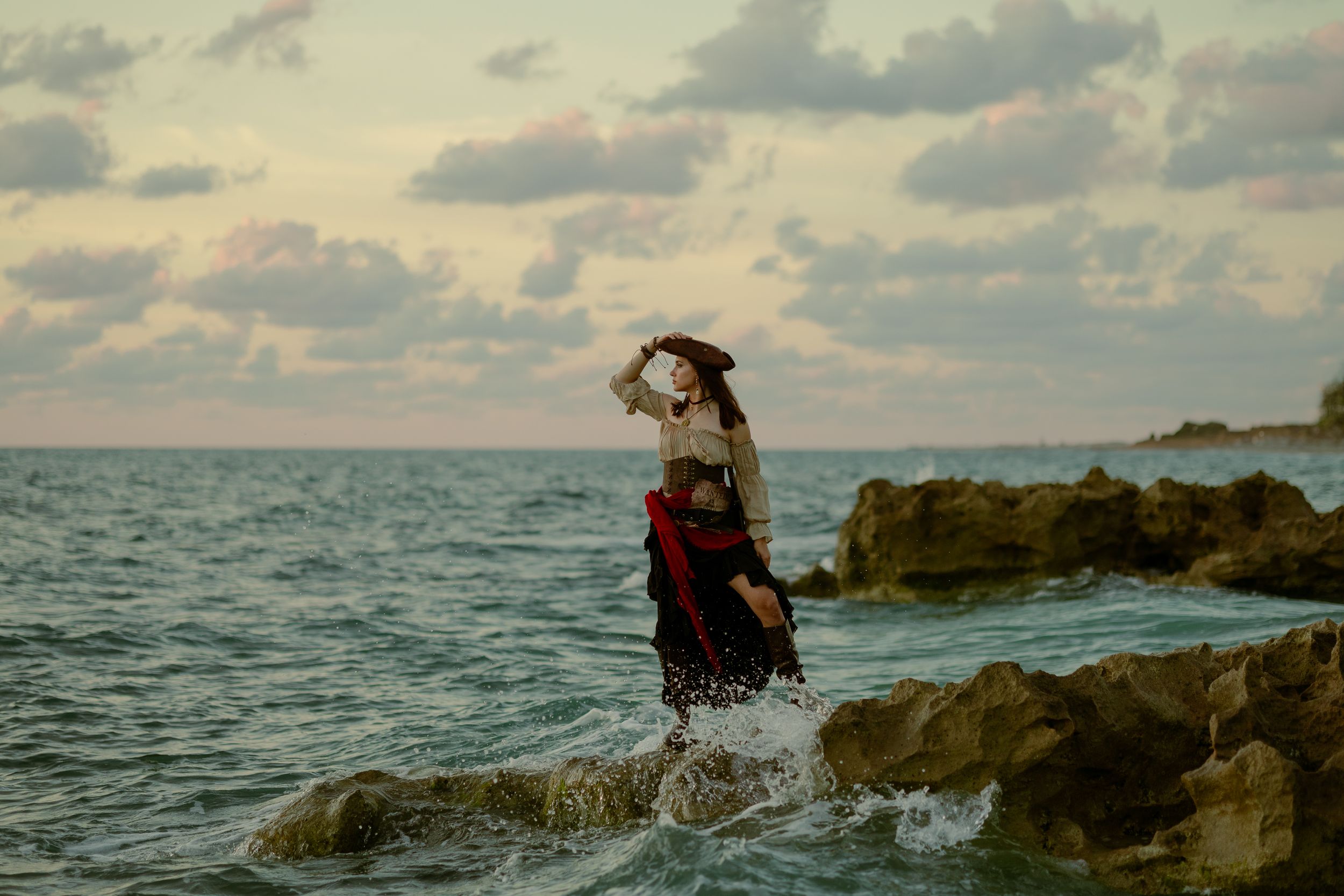 Pirate woman stands on rocks at sunset, waves crashing behind her. Cinematic cosplay editorial in South Florida by Mannyportraits.