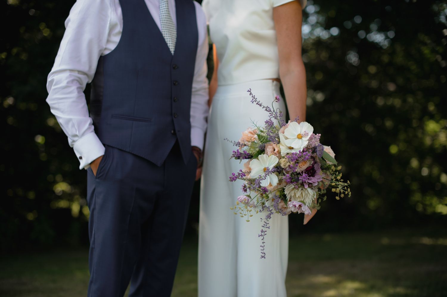 Elegant wedding couple embracing outdoors in formal attire with floral bouquet.