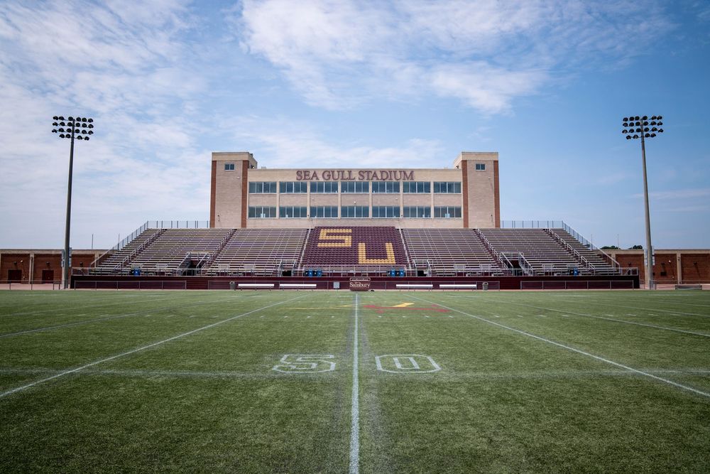 An empty high school football stadium with brick buildings in the background under cloudy skies.