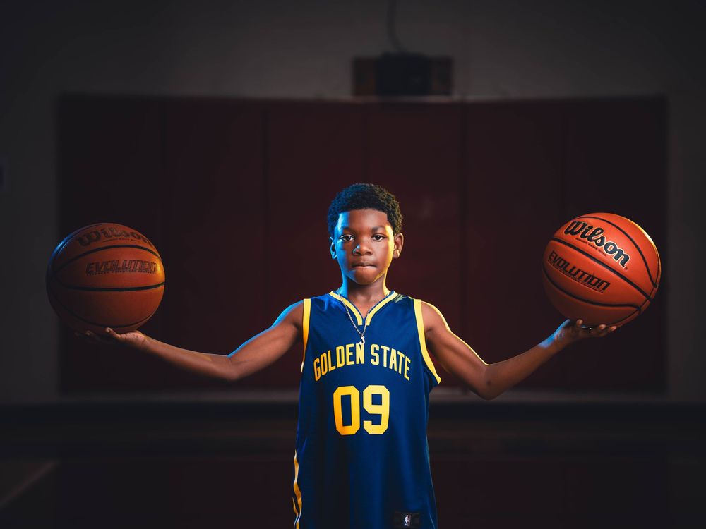 Basketball player in blue and gold uniform holds two basketballs with arms outstretched in dramatic lighting.