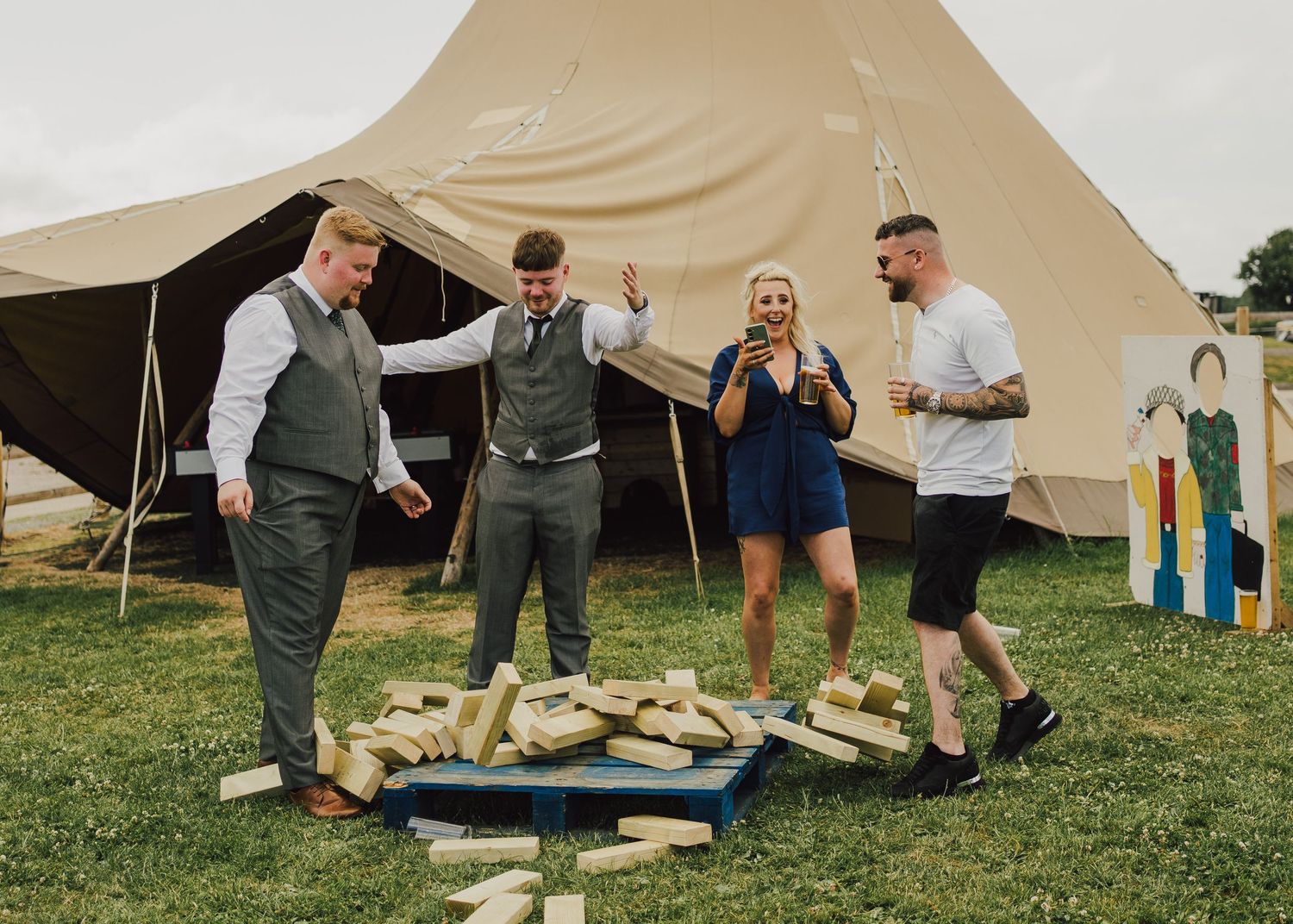 A group plays a garden game with wooden blocks on grass in front of a large beige tent at an outdoor event.