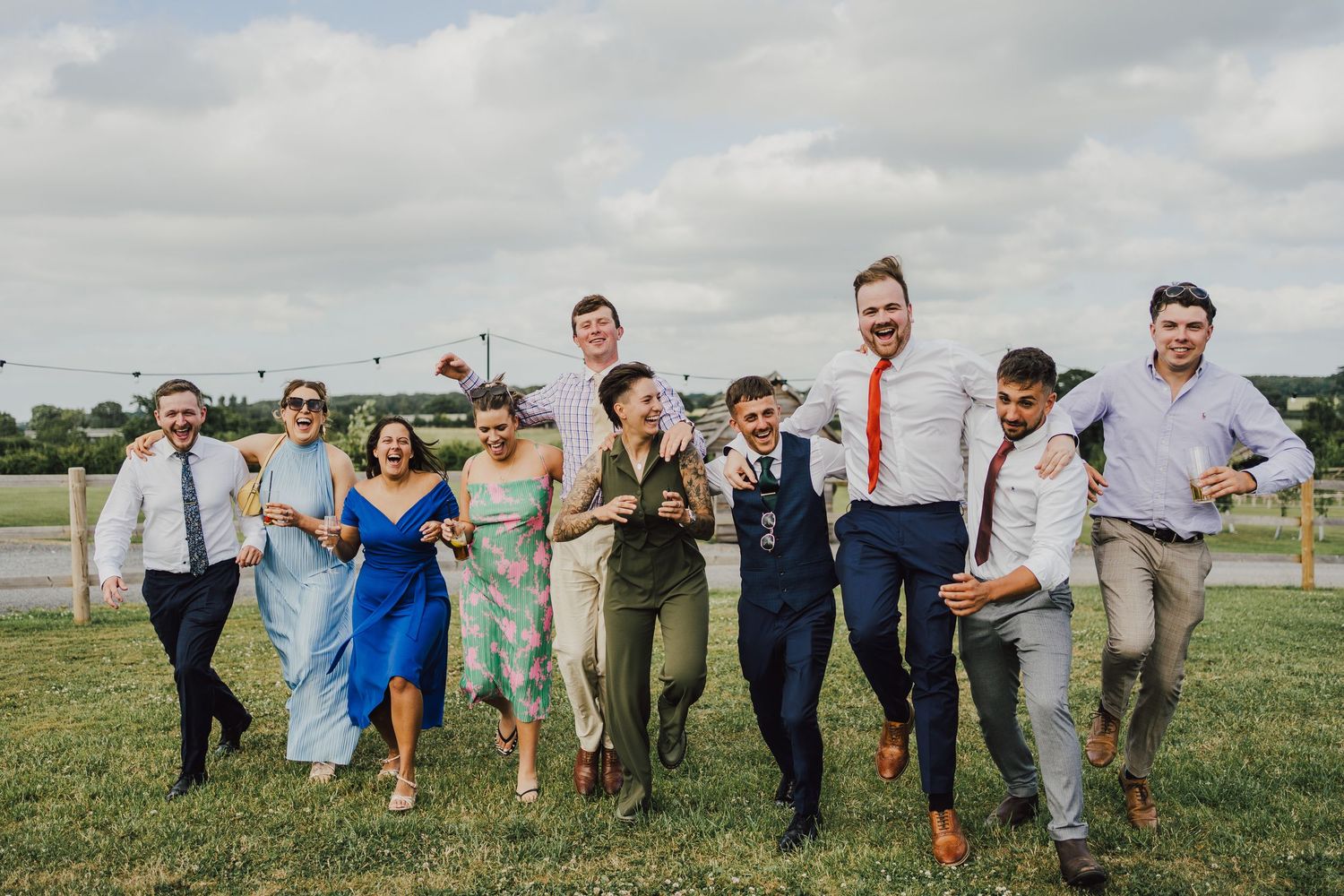 A group of wedding party members playfully poses in a grassy field under a cloudy sky.