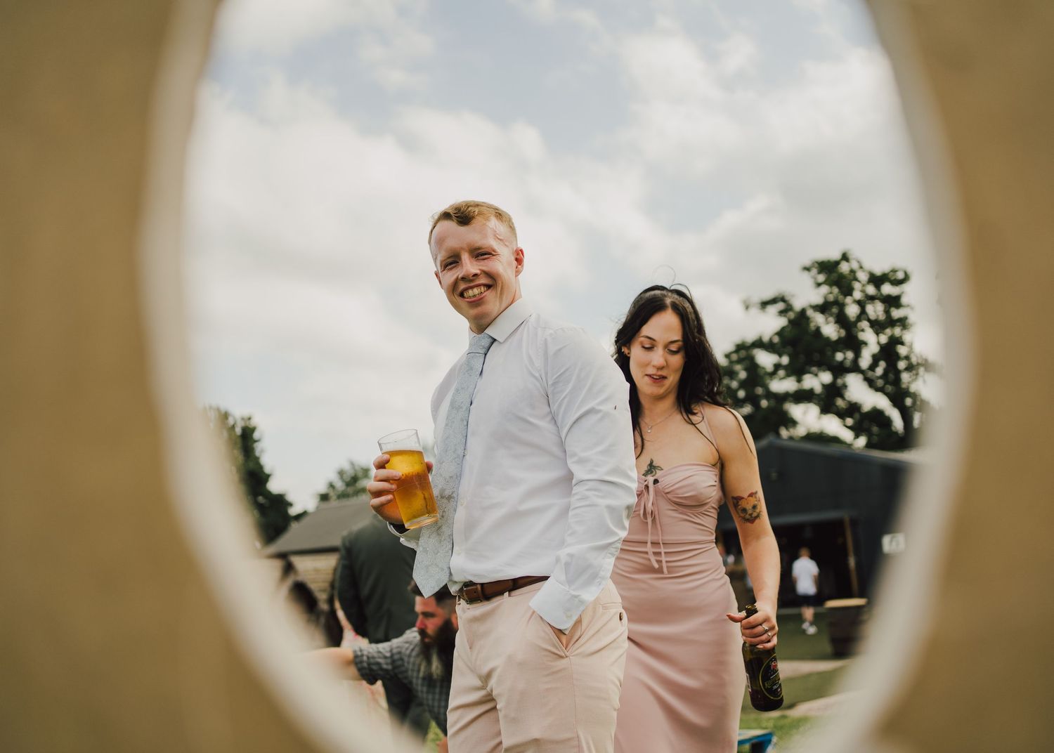 A couple in wedding attire walks together outdoors through a circular frame, with the person holding a beverage.