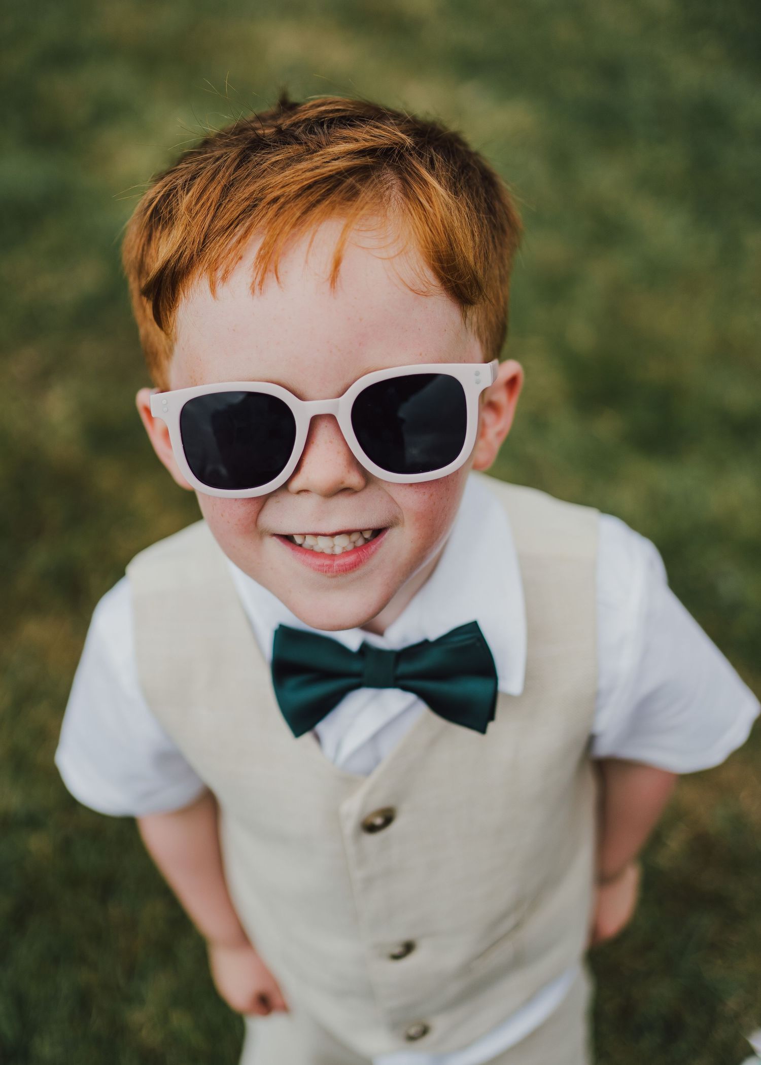 A child in a white vest and bow tie makes fun expressions through sunglasses in a sequence of outdoor photos.