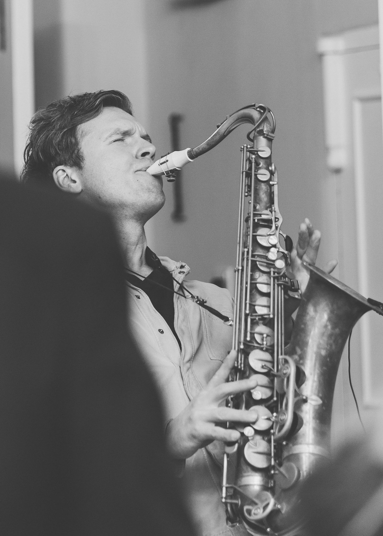 A musician plays a saxophone in black and white, captured in an intimate close-up performance shot.