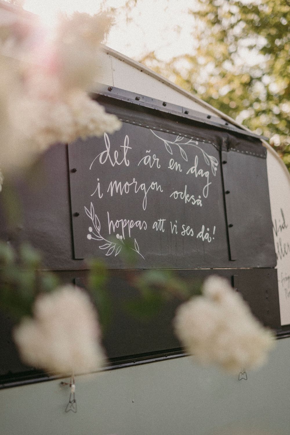 Handwritten text in white chalk on black metal box with blurred flowers in foreground.