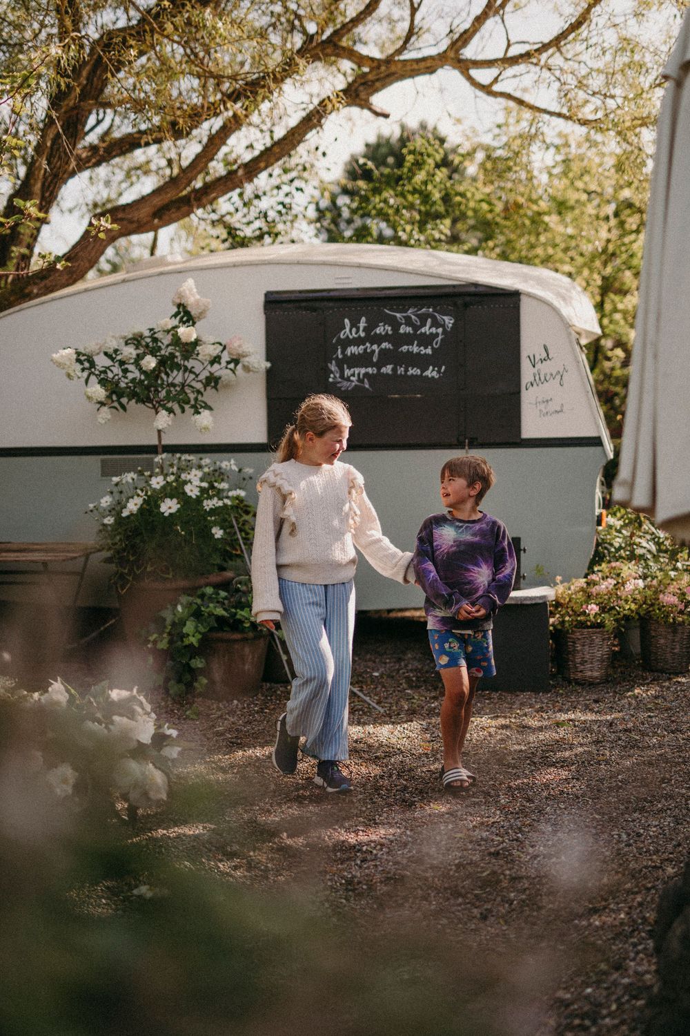 Two children walking together in front of a white camper trailer on a sunny autumn day amid trees and foliage.