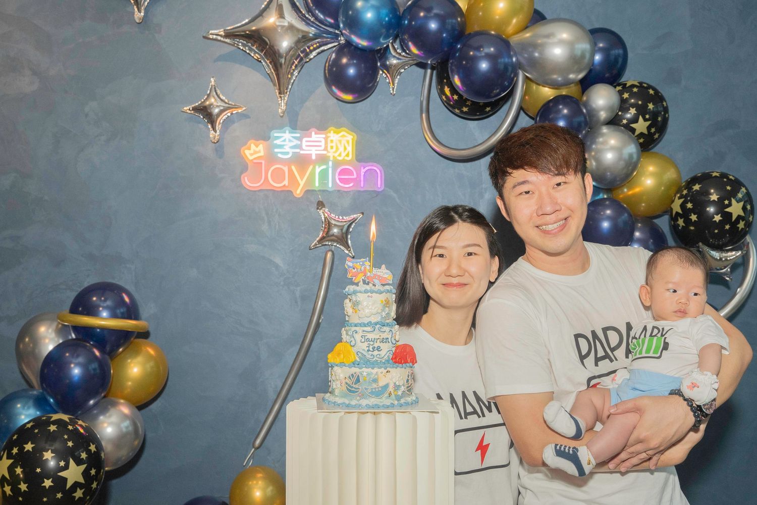 Birthday celebration setup with blue, gold, and silver balloons arranged against a grey wall, featuring star decorations.