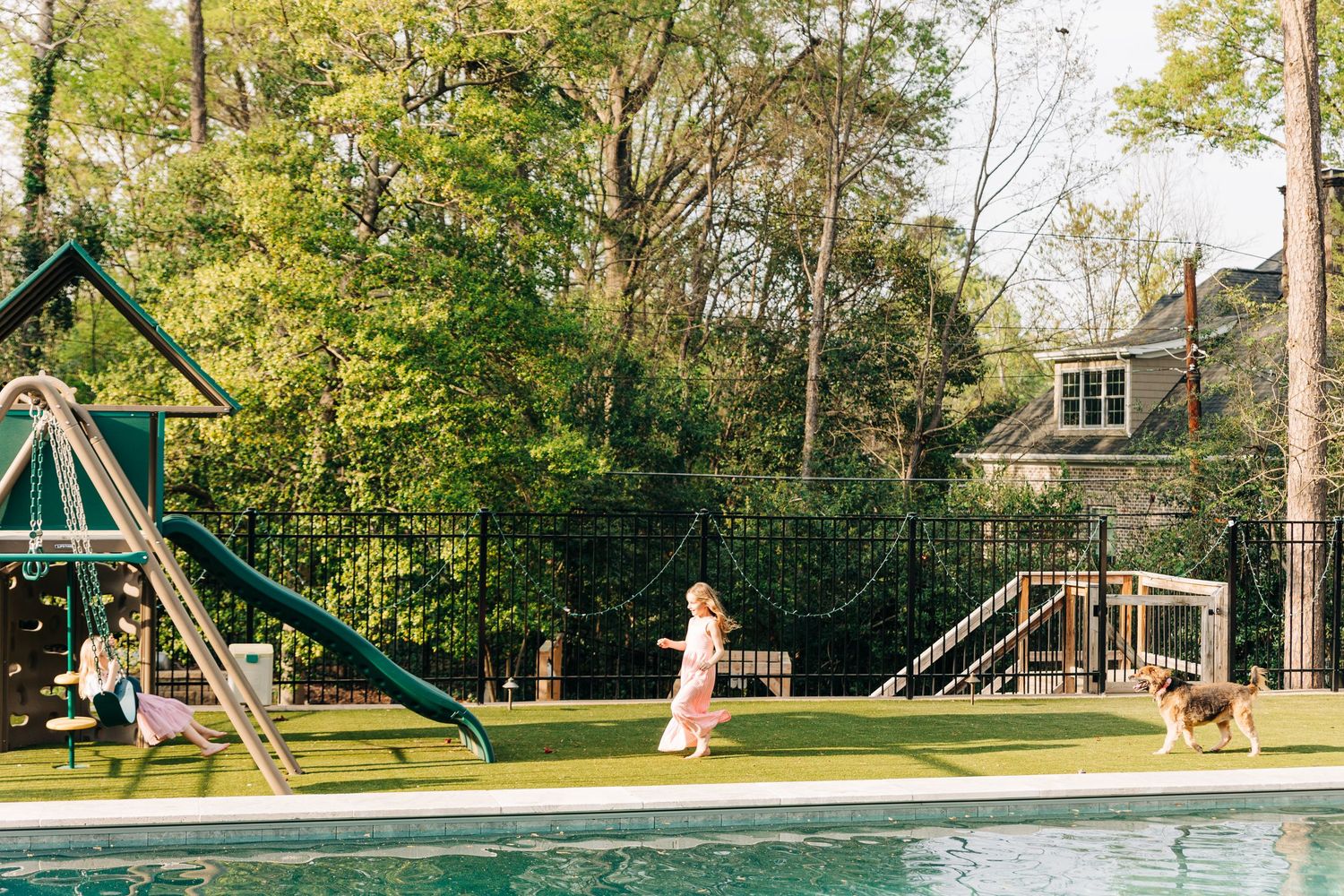 Panoramic view of a modern backyard playground with swings and slide set against lush green trees and landscaping.