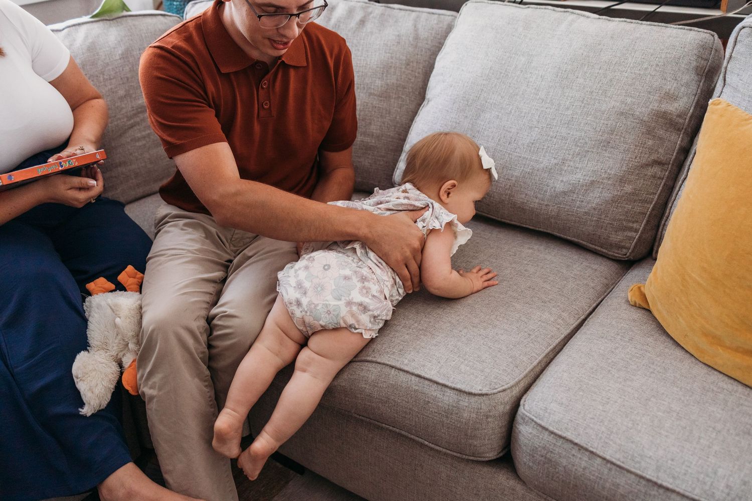 A baby wearing a patterned onesie lies on a gray couch while being comforted by someone in a brown shirt.