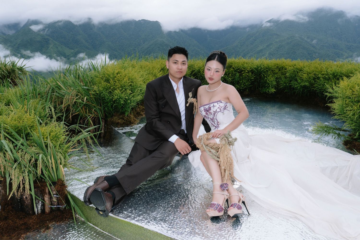 Wedding couple sits together on reflective rice terrace path with lush green mountain backdrop.