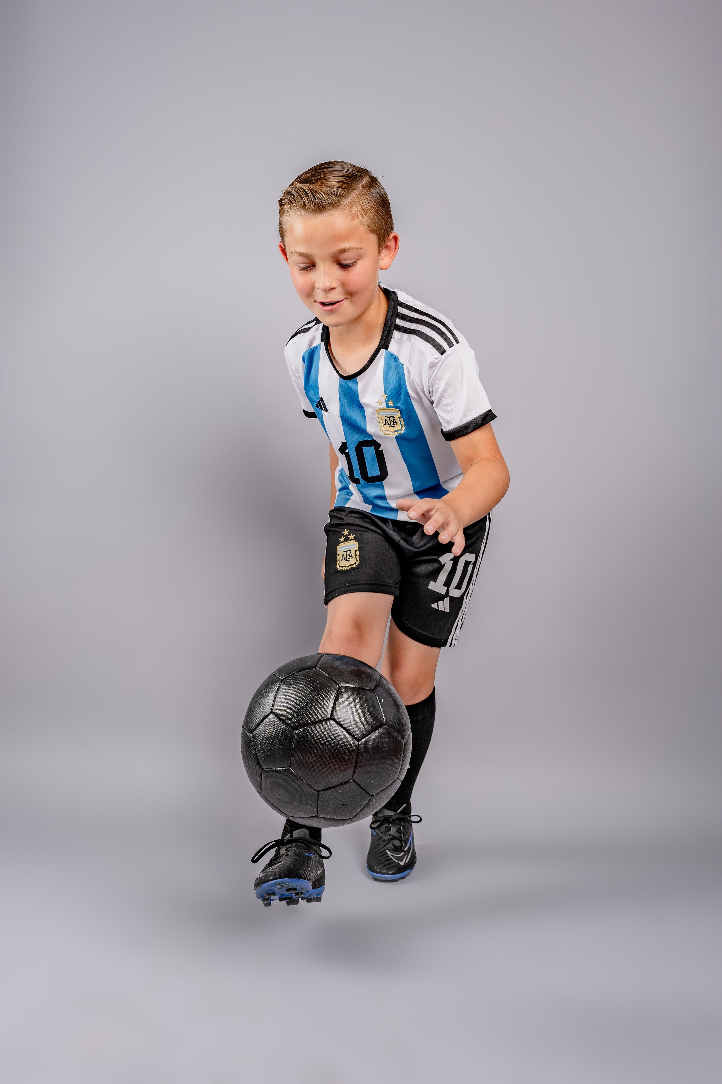 Young athlete in Argentina soccer jersey performs tricks with a soccer ball in studio against gray background.