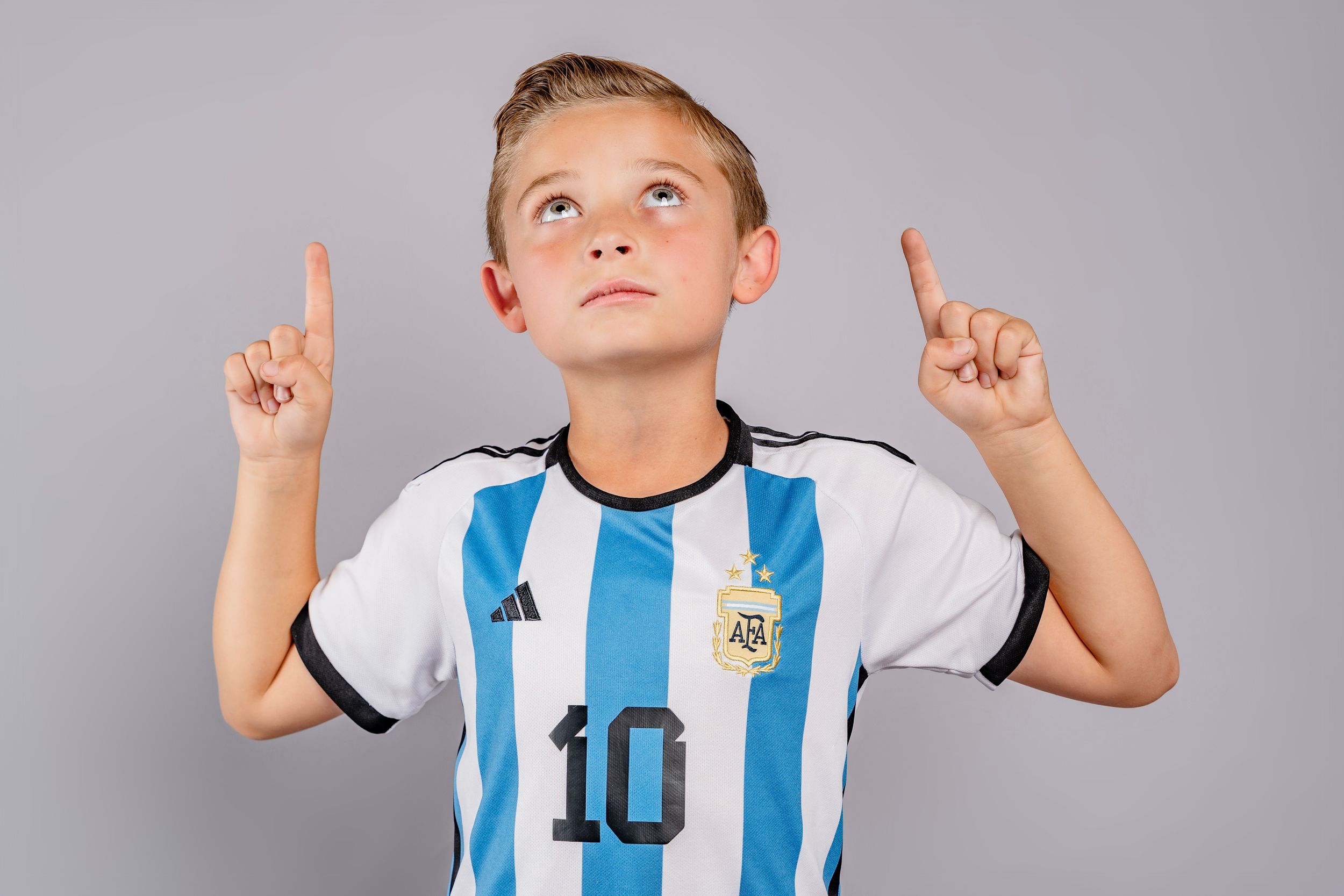 Soccer player in blue and white striped jersey points upward while posing against a gray background.