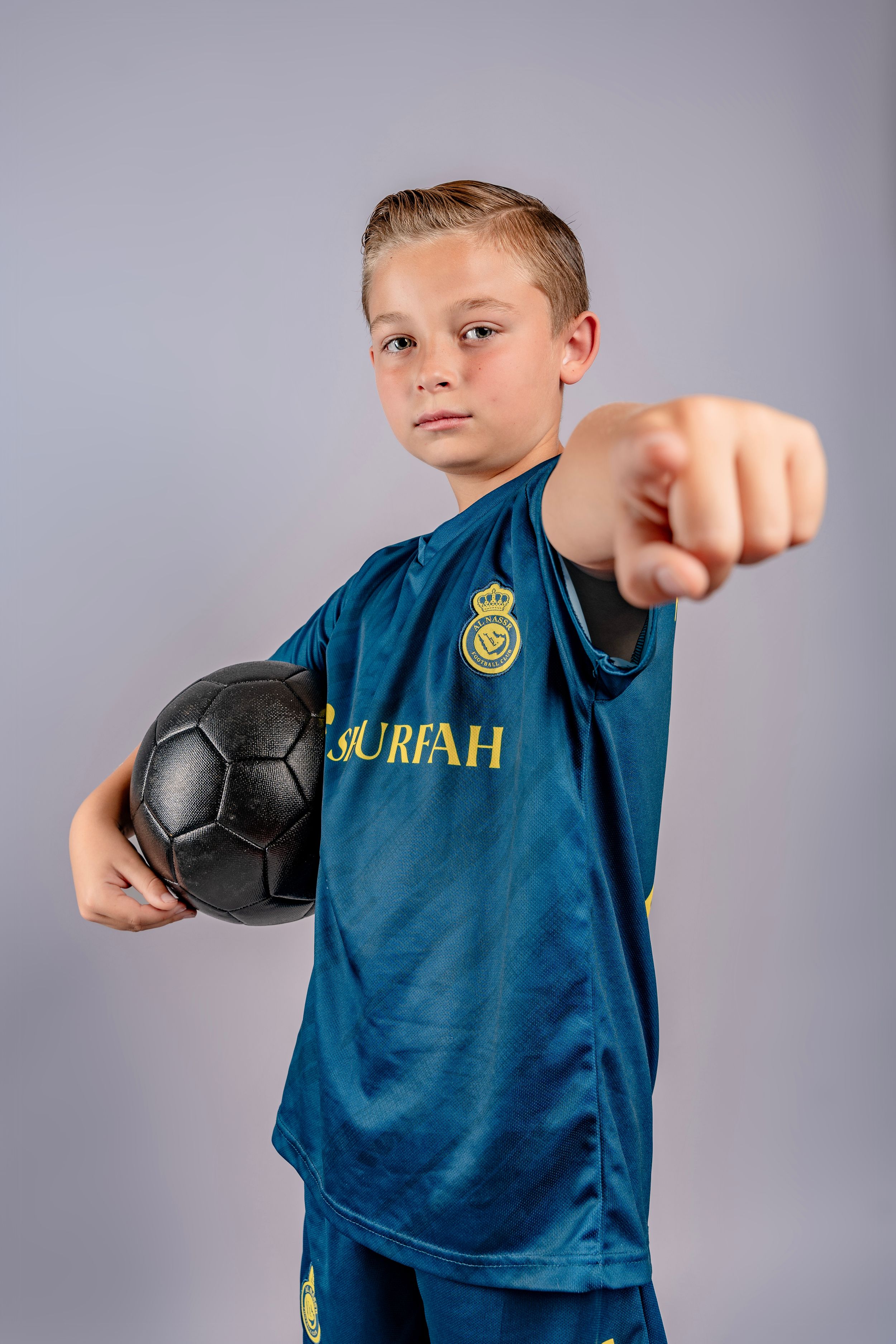 Young soccer player wearing blue jersey demonstrates striking poses with black ball against gray background.