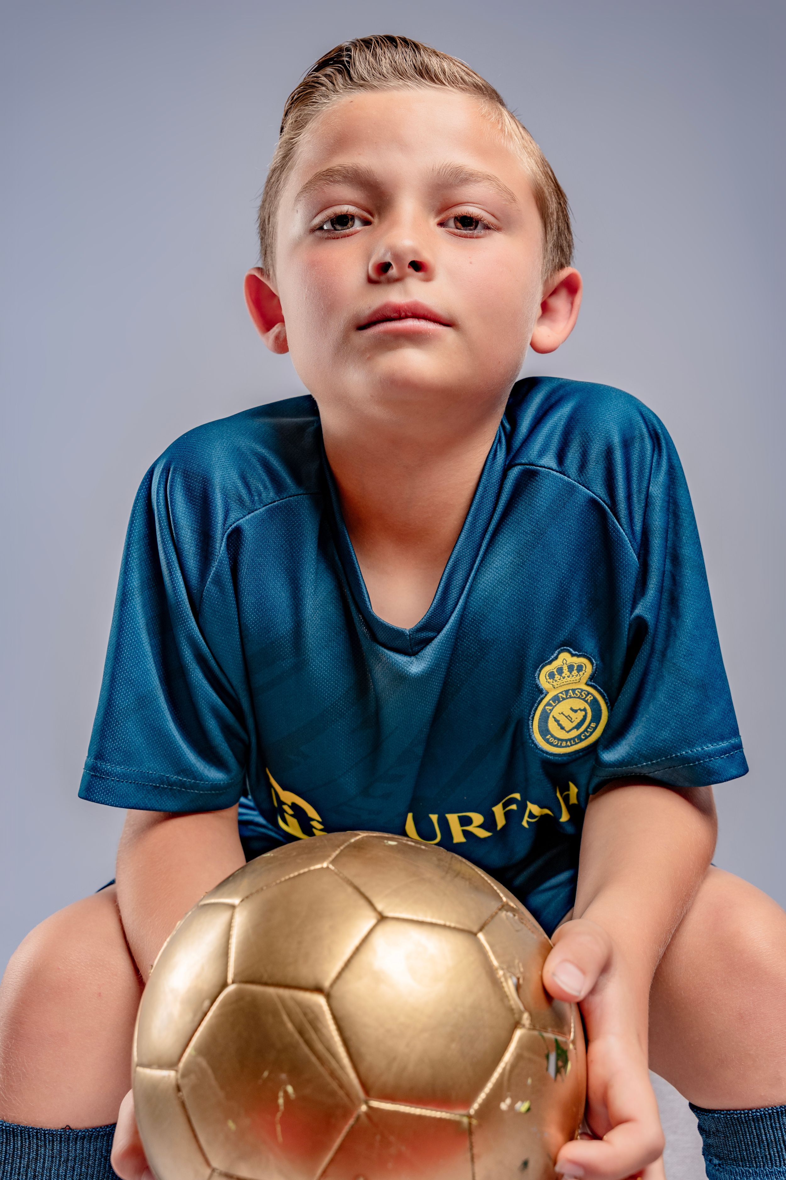 Young athlete in a navy blue sports jersey poses playfully with a shiny gold soccer ball in a series of shots.
