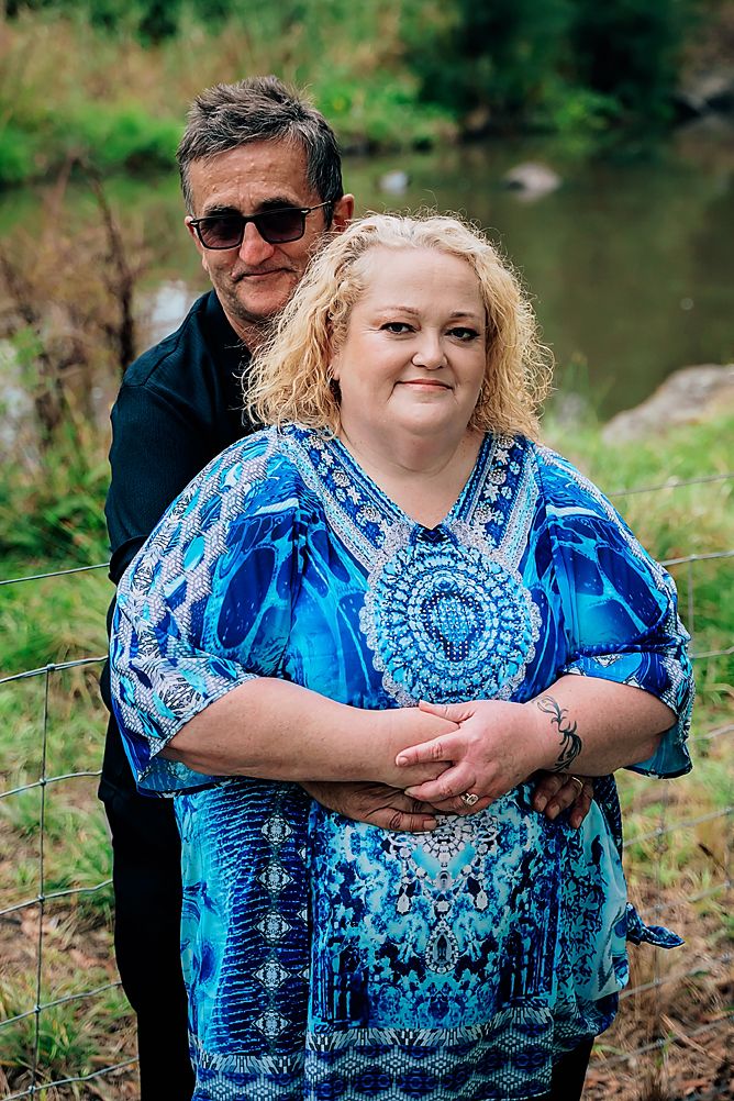 A couple in casual attire embraces by a scenic pond with trees and greenery in the background.