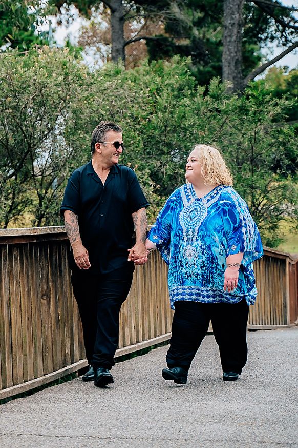 Two people stroll together on a wooden bridge walkway dressed in contrasting black and blue outfits on a sunny day.