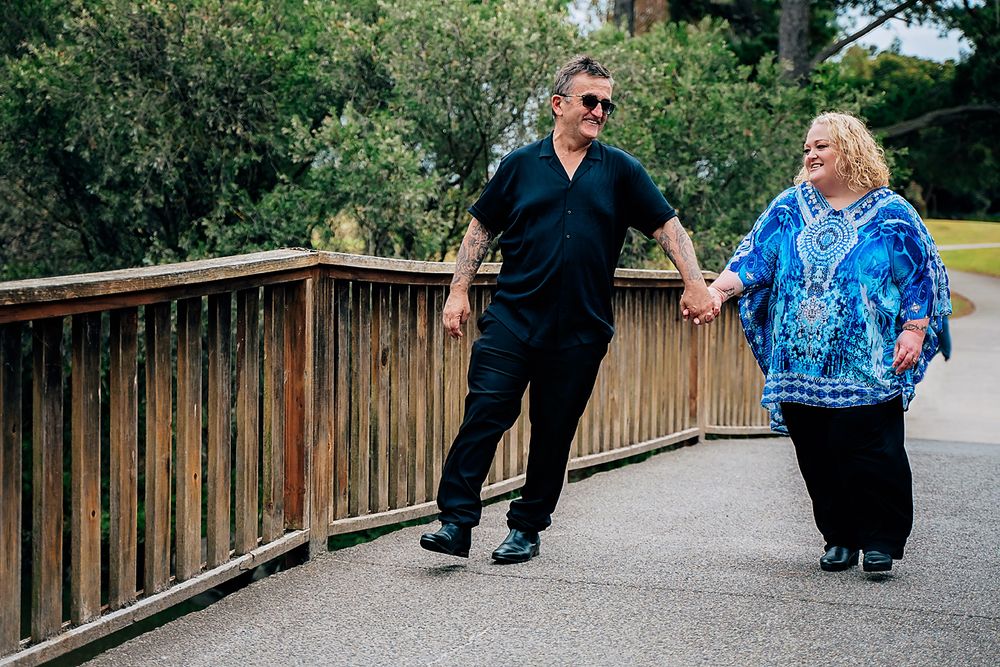 A couple walks hand in hand on a wooden boardwalk through a scenic park area in summer.
