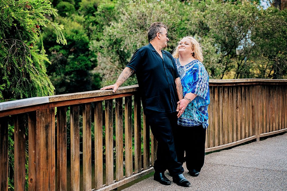 Couple sits together on wooden log benches during outdoor engagement photo session in lush green park setting.