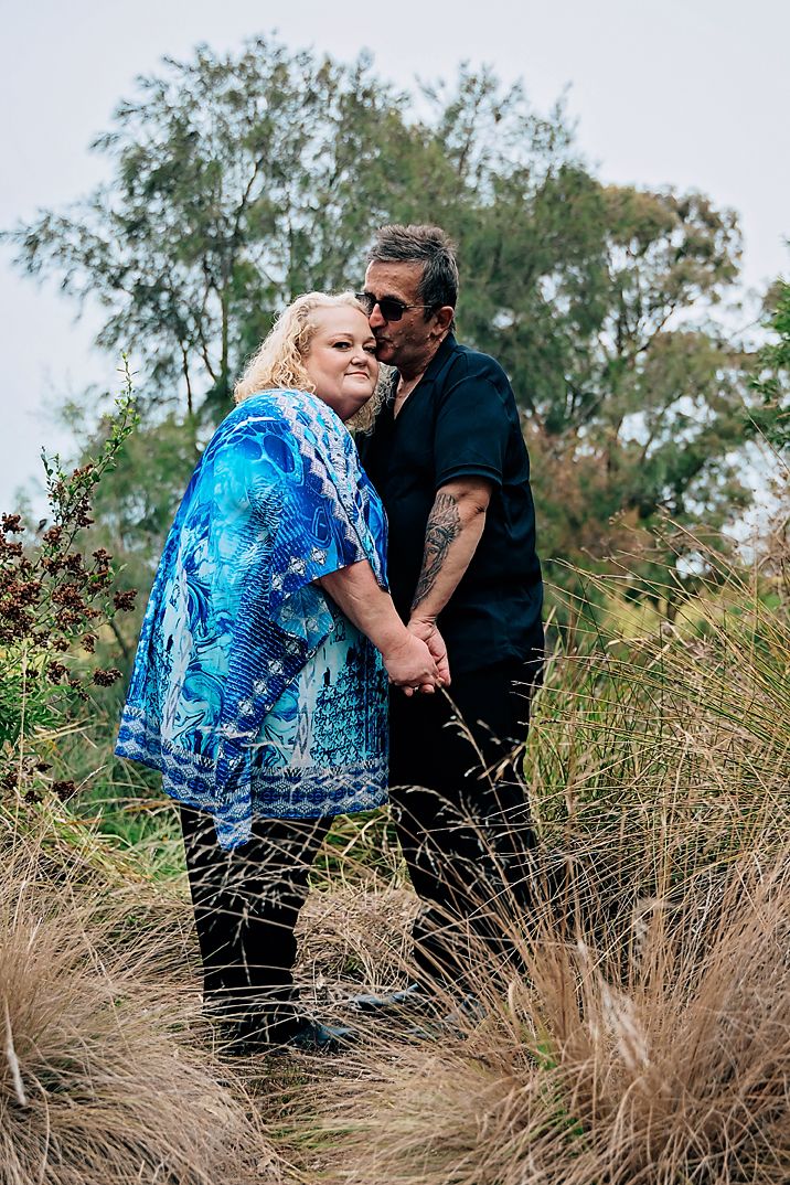 A couple embraces in tall grass on a sunny day, with the person wearing a flowing blue patterned dress.