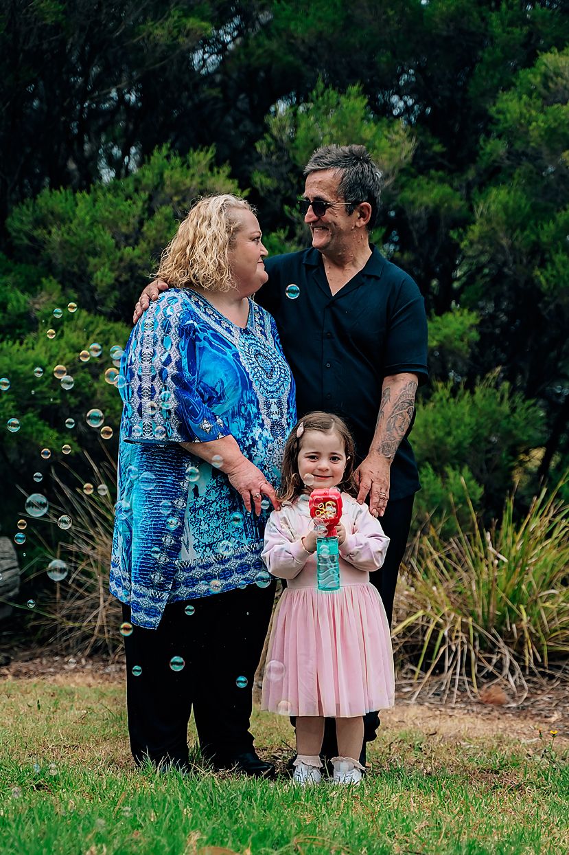 Family plays together on grassy lawn with bubbles floating in the air and ornamental grasses in background.