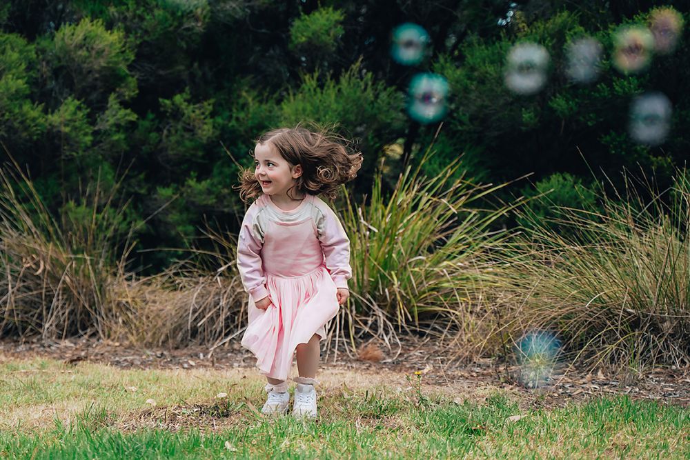 A child in a pink dress walks through an outdoor setting with soap bubbles floating in the air.