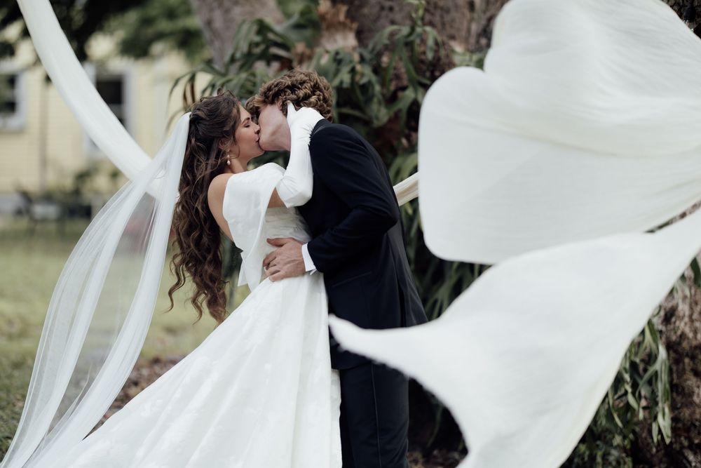 A bride and groom share a romantic kiss as flowing white veils float in the breeze during their outdoor wedding.