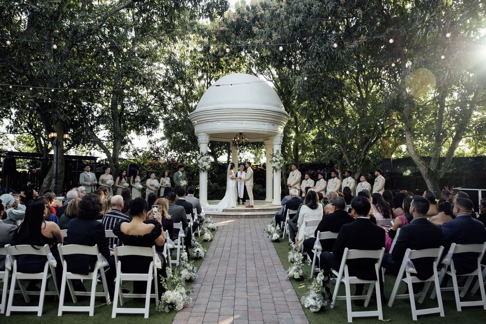 White chairs line both sides of an outdoor garden pathway leading to a white domed gazebo for a wedding ceremony.