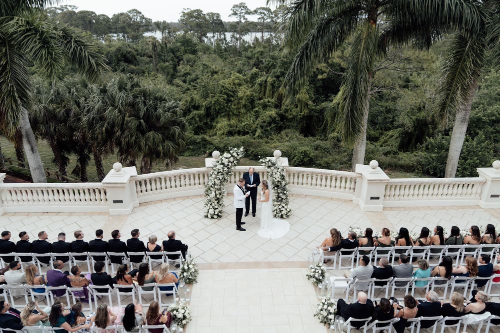 Guests seated in white chairs at an outdoor wedding ceremony on a stone terrace surrounded by palm trees and greenery.