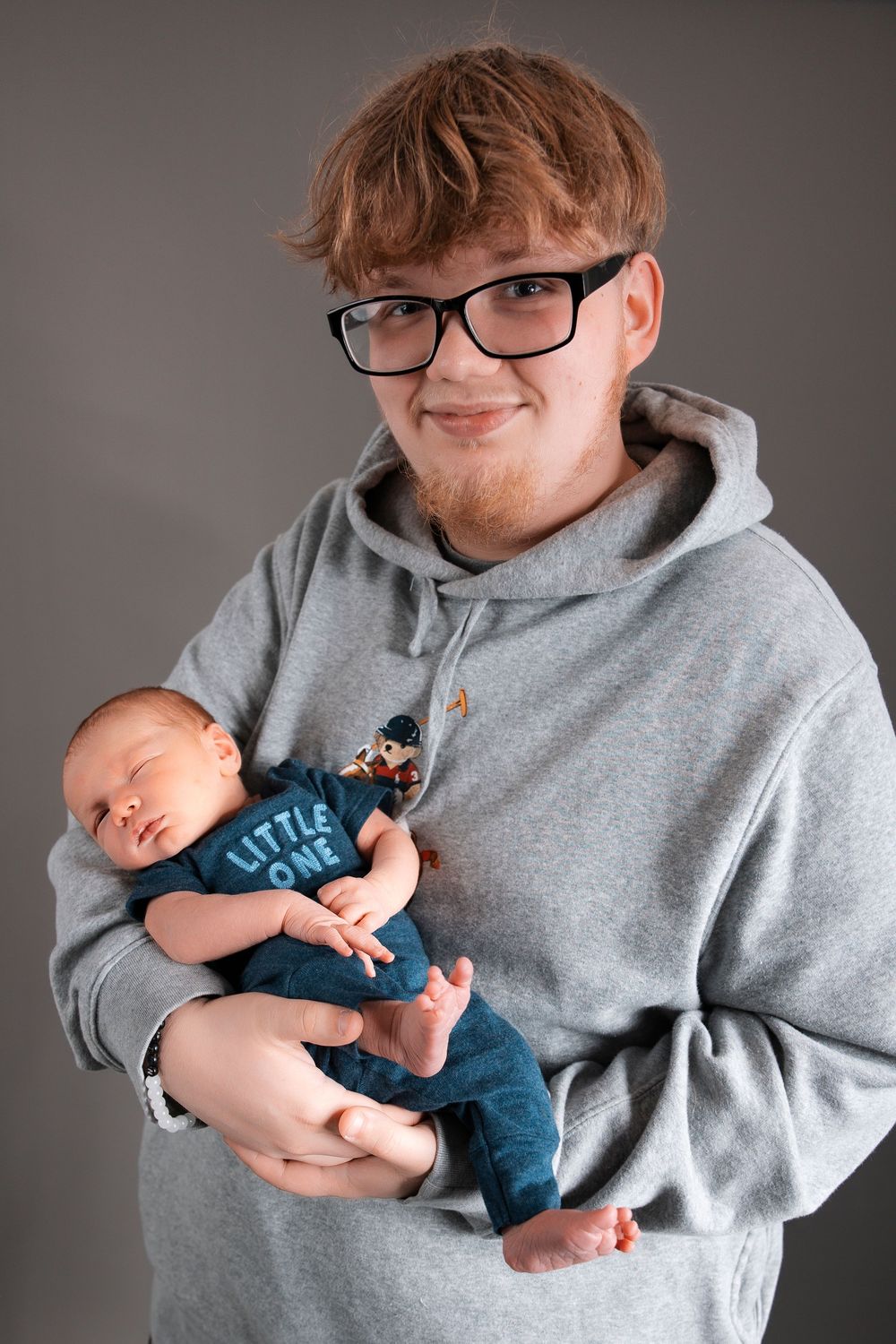 Person wearing gray hoodie and glasses tenderly holding sleeping newborn baby in blue onesie.