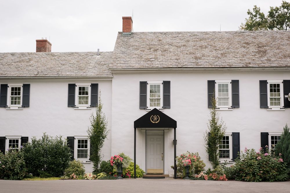 Long white colonial style building with black shutters and multiple doorways adorned with flower beds.