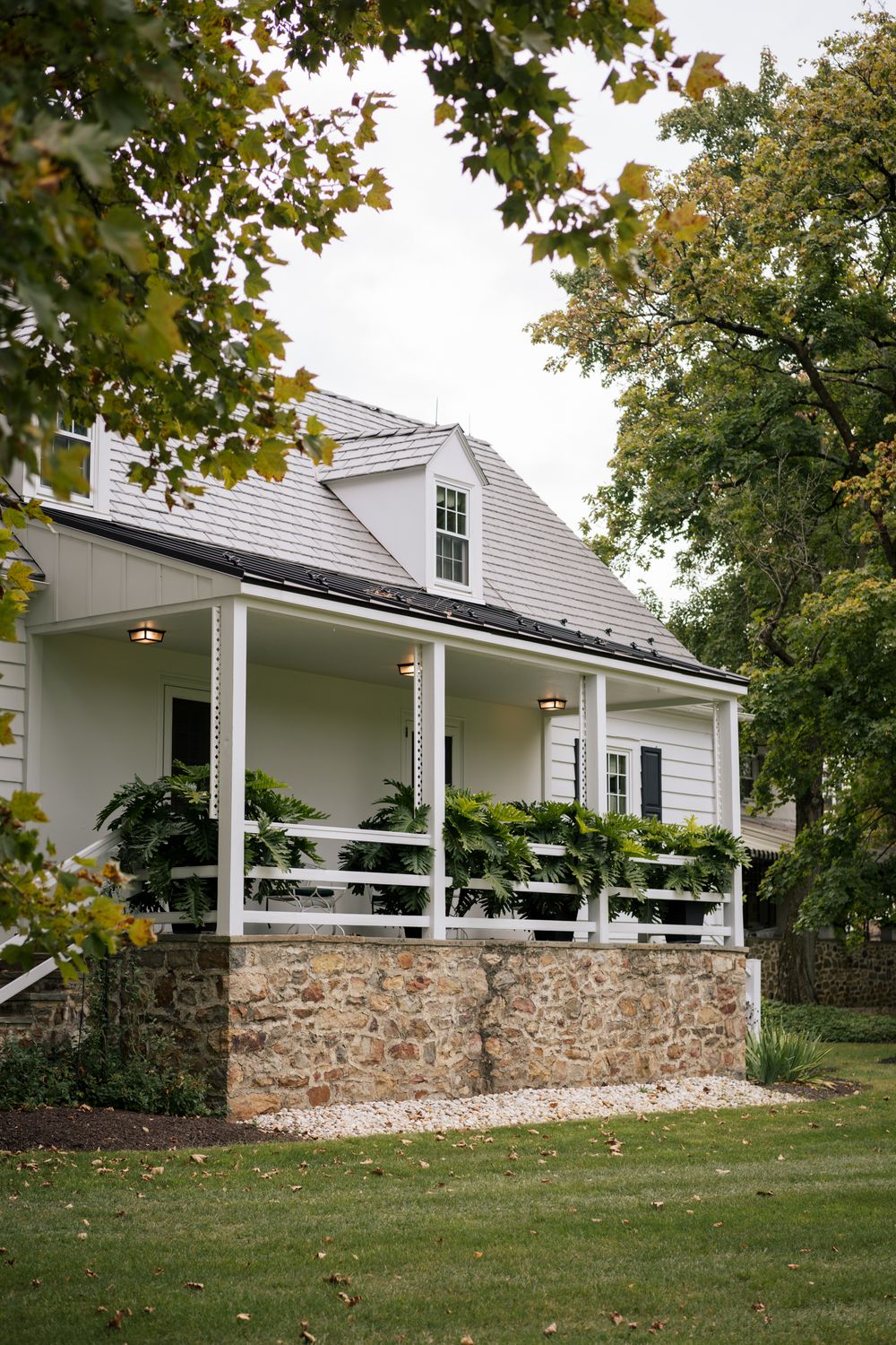 White farmhouse with wraparound porch and stone foundation sits nestled among autumn trees.