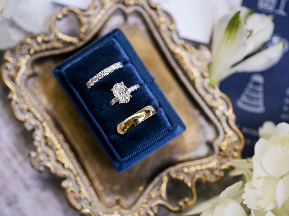 Elegant engagement rings rest in blue velvet ring boxes with ornate gold trim and white flowers.