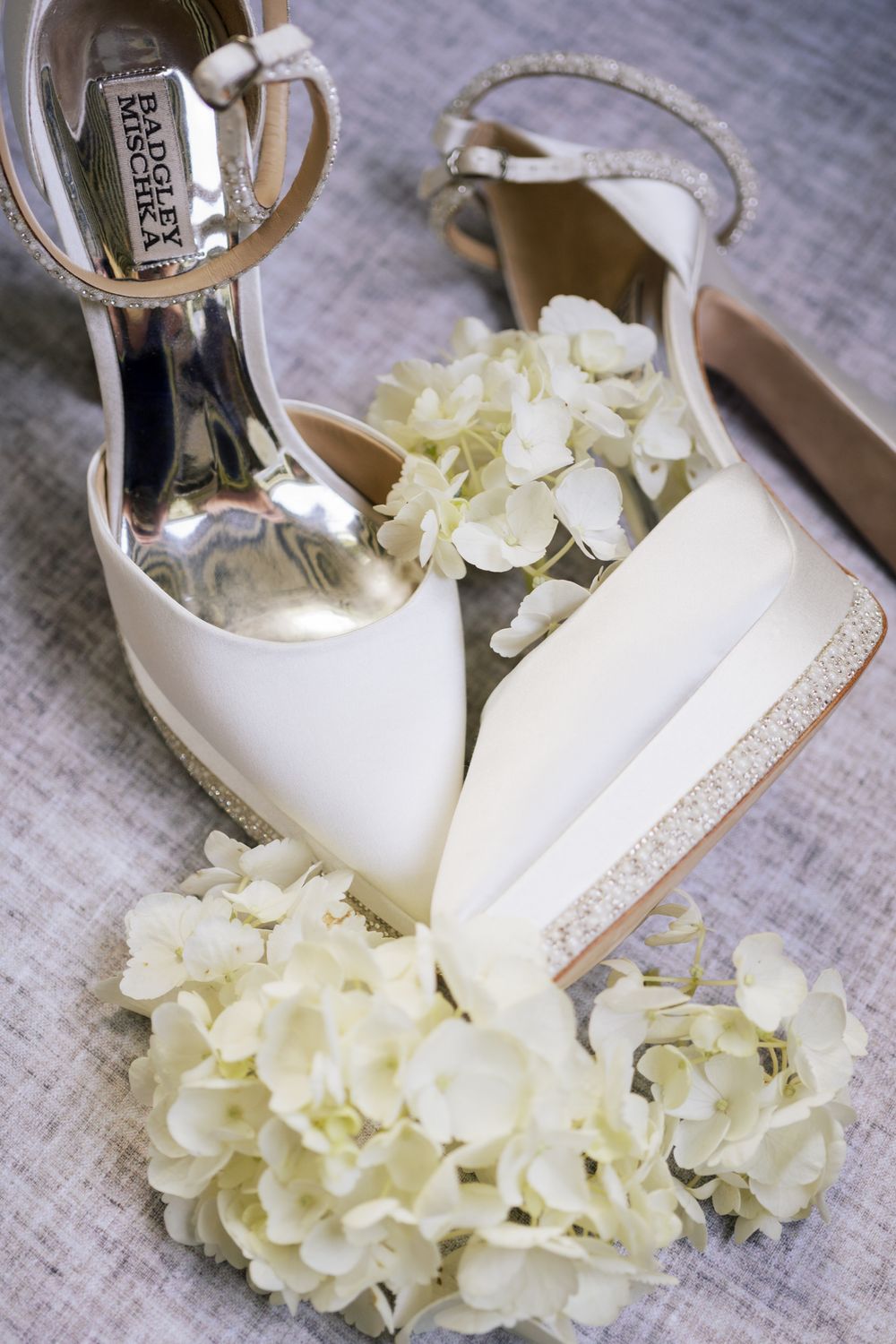 White bridal heels placed next to delicate cream hydrangea flowers on a rustic surface.