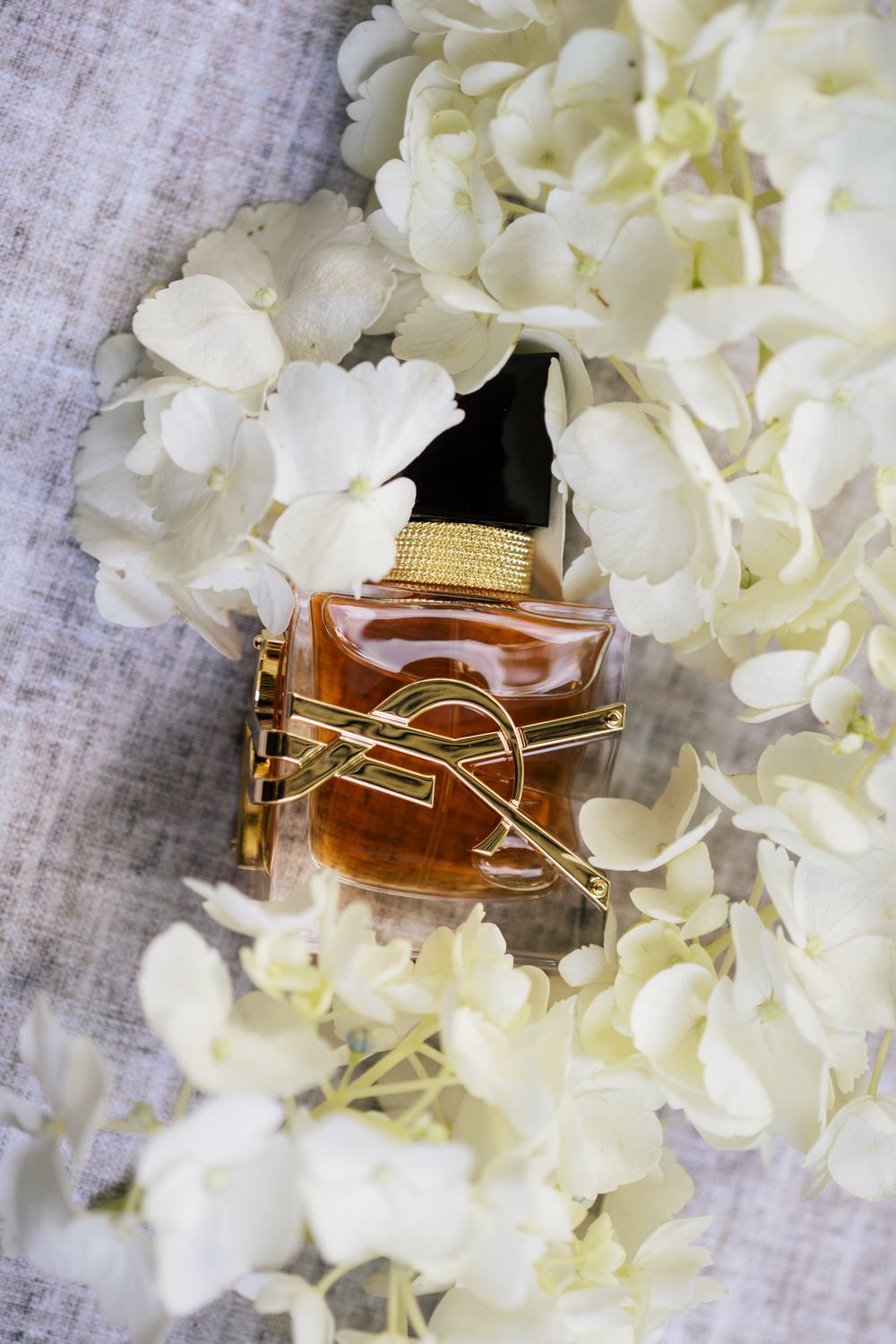 A perfume bottle nestled among delicate white hydrangea flowers on textured fabric.